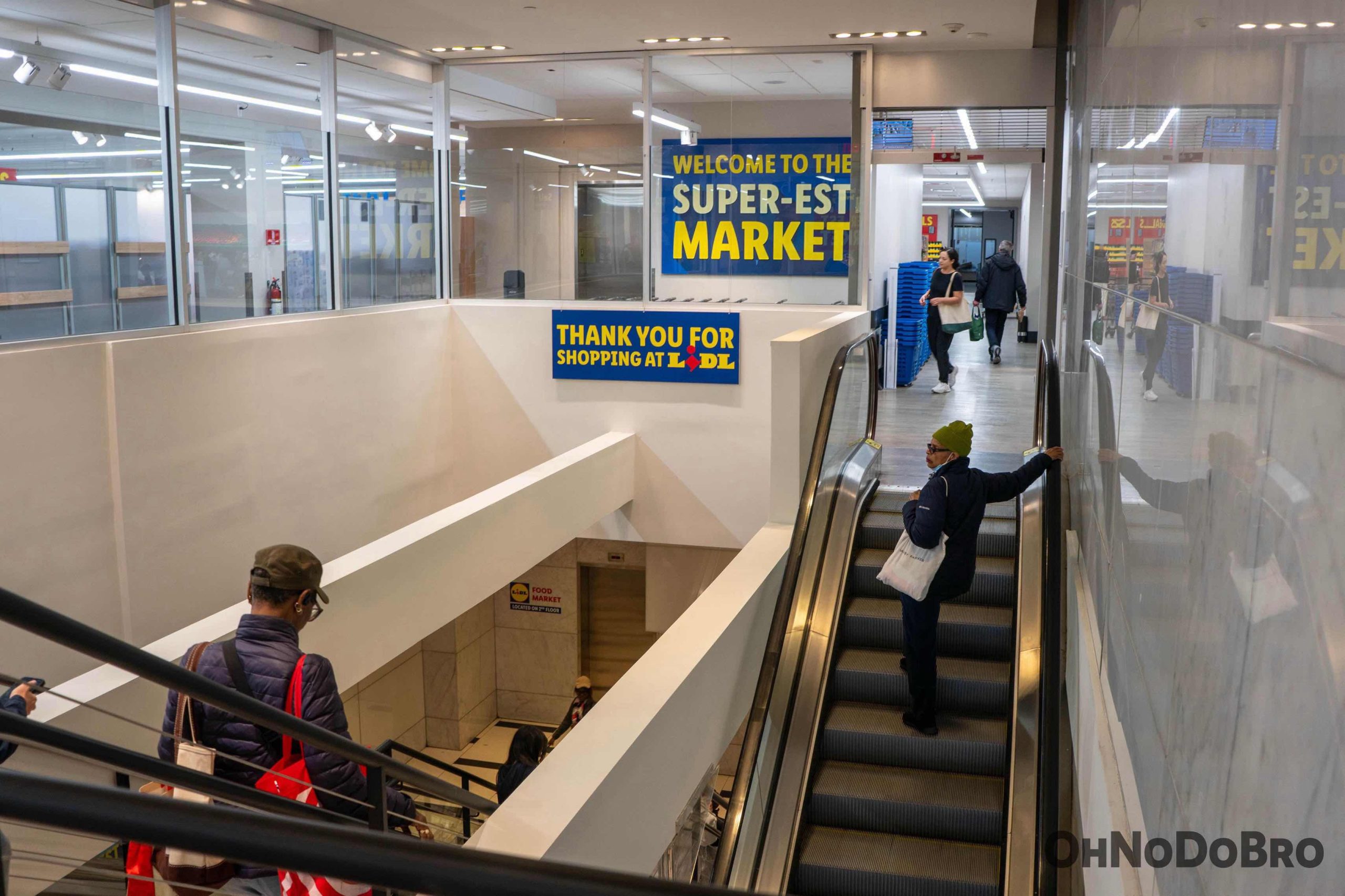 Exiting Lidl down the escalator. You can see both the up and down escalators, as well as a sign that says "welcome to the super-est market" and "thank you for shopping at lidl"